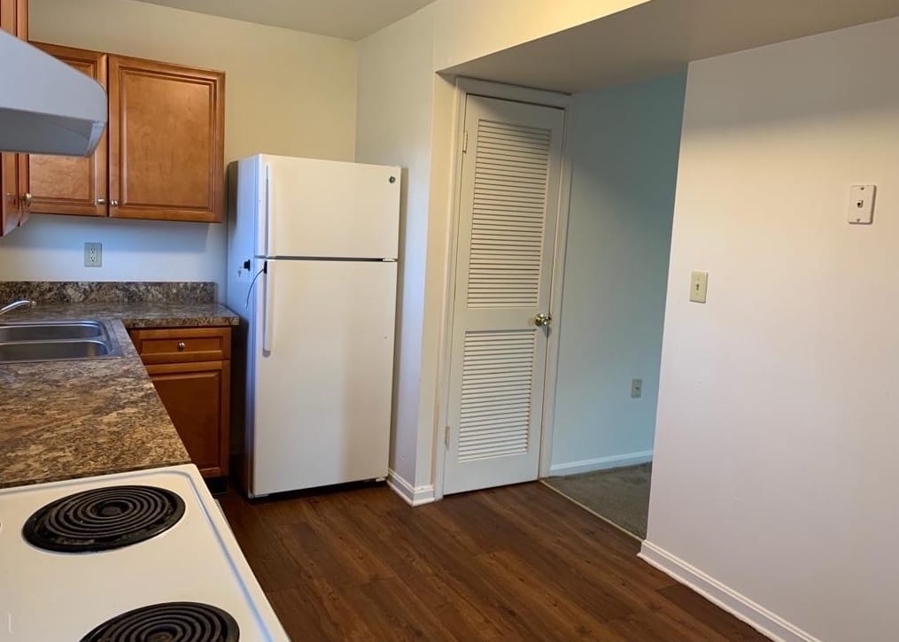 A Thurmont Village kitchen with a white refrigerator and a louvered pantry door, opening toward the carpeted living area