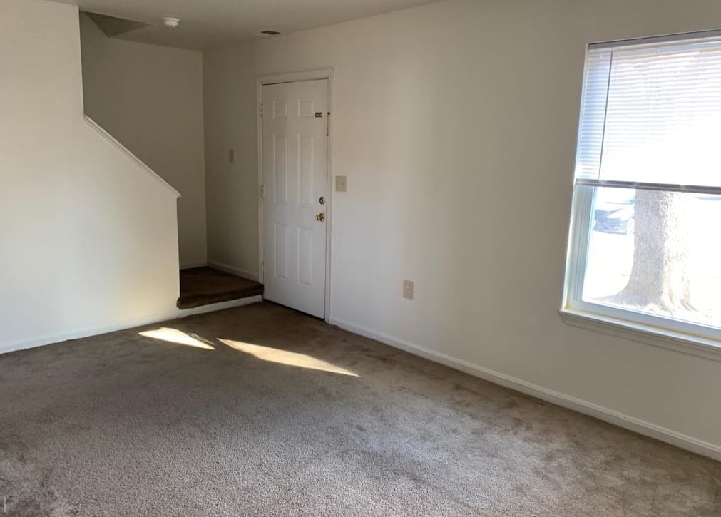 The carpeted living room in a Thurmont Village townhouse, with the front entry door and the staircase up to the bedrooms